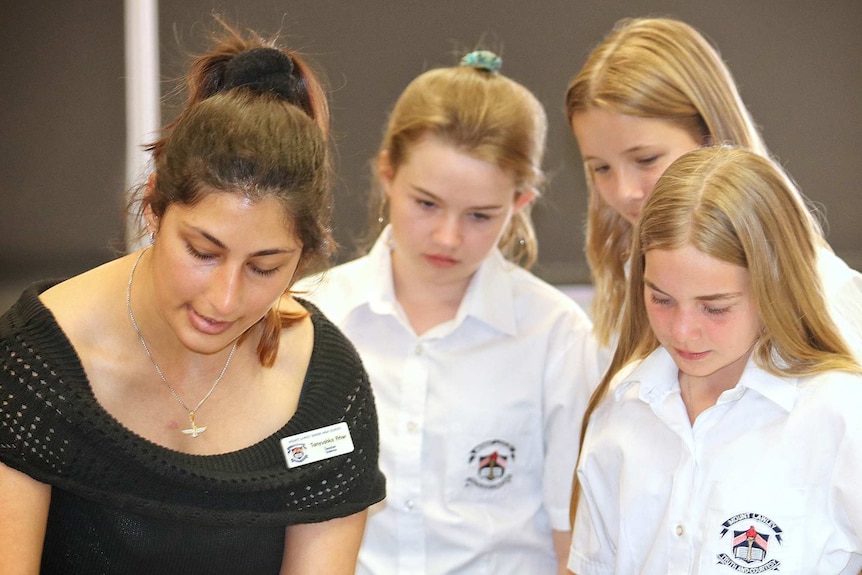 A female school teacher stands with three female Year 7 students looking down at something.