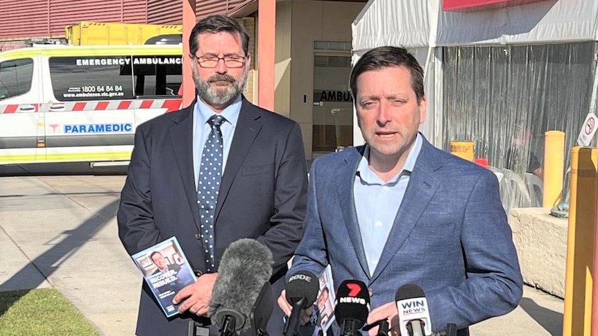 Two men in suits talking into a bank of microphones outside a hospital.
