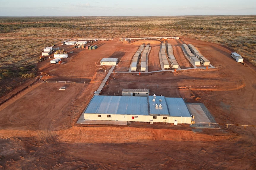 Mining village in the foreground of a vast, shrub-based landscape, with red earth scattered between portable buildings.