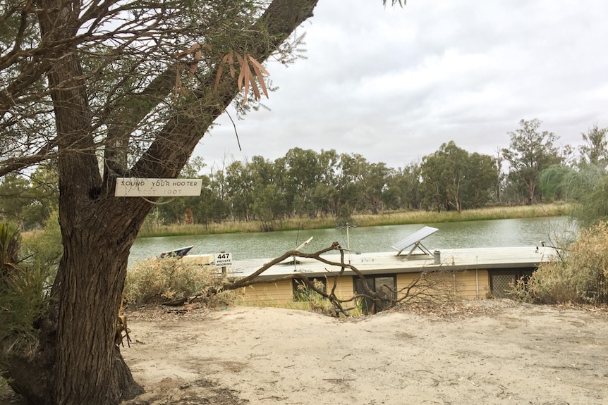 An old houseboat, partially seen above the level of the river bank, with a sign on a tree that reads Sound Your Hooter