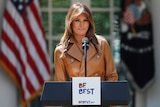 Melania Trump smiles, standing behind a lectern with the slogan Be Best written on it, with american flags behind her