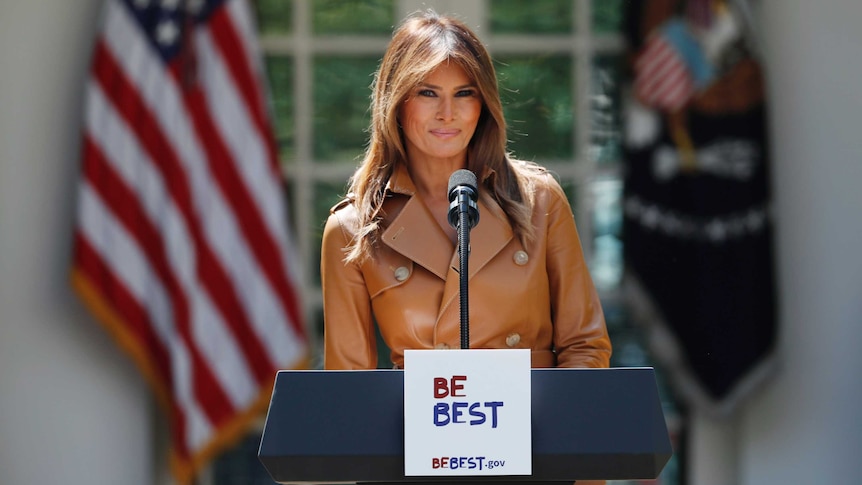 Melania Trump smiles, standing behind a lectern with the slogan Be Best written on it, with american flags behind her