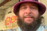 A man with a red hat and bushy beard smiles at the camera. He is wearing a bright shirt.