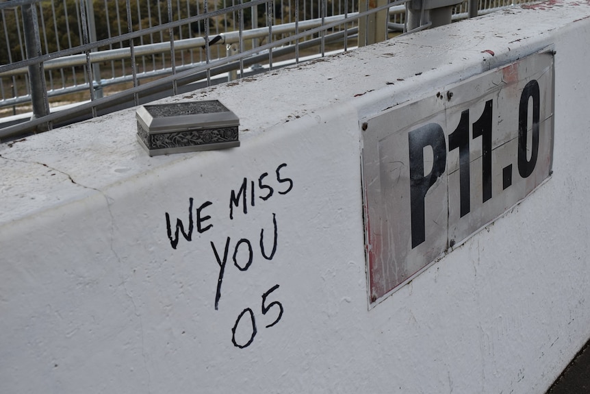 A white dividing wall with "We miss you 05" written on it with an ornate urn containing his ashes sitting on the wall.