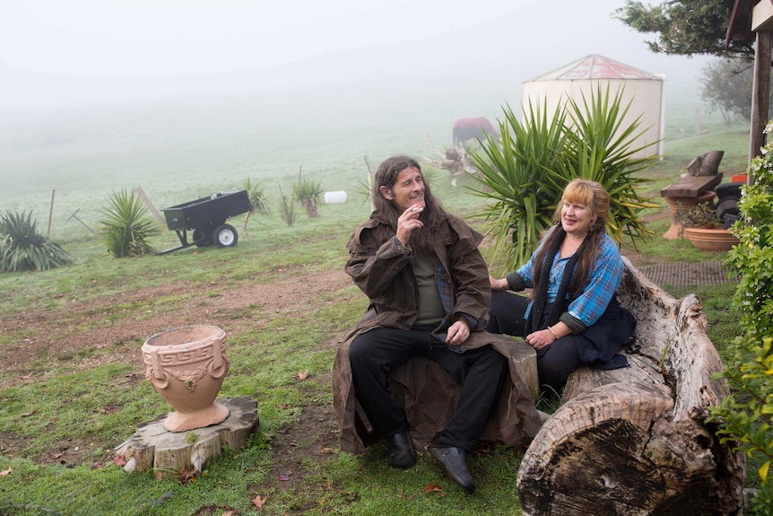 A man and a woman sit on logs in the backyard of a large property with dew on the grass surrounding them