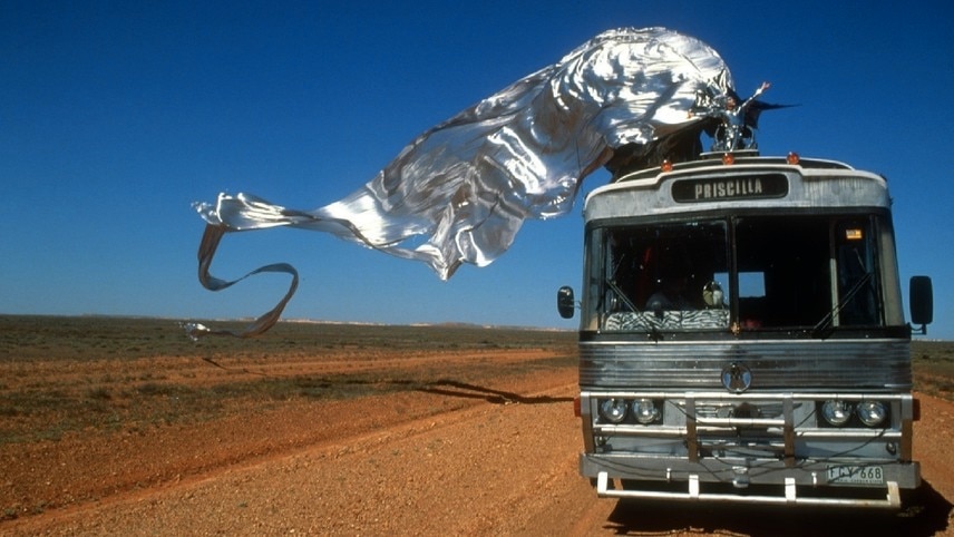 A silver bus travels along a dirt road, as a performer in a long flowing silver gown stands atop it. 