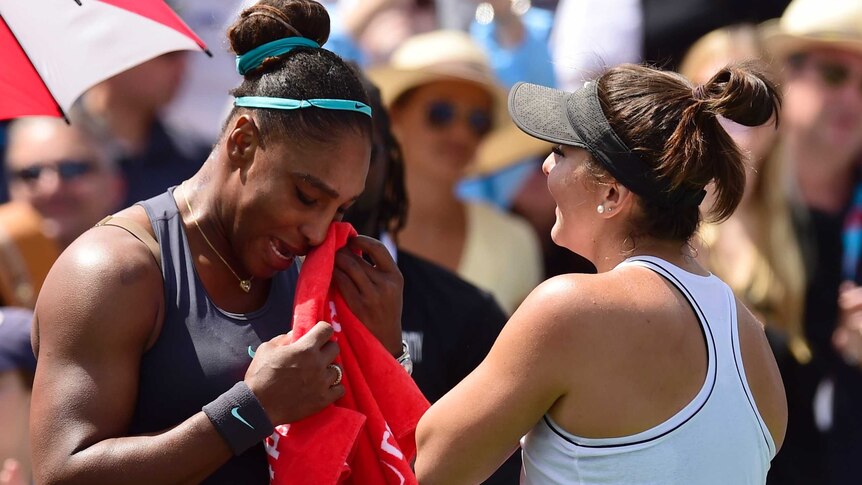 Serena Williams holds a towel to her face while speaking to Bianca Andreescu on a tennis court.