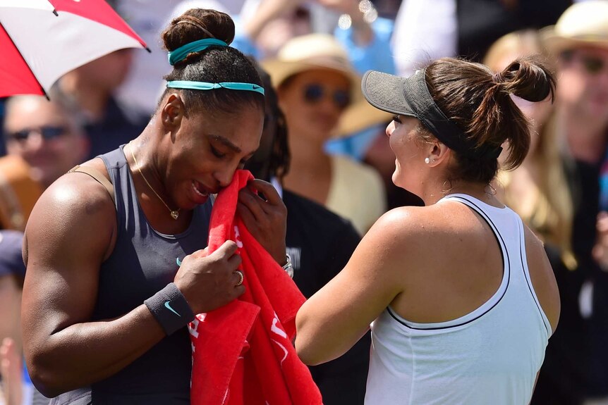 Serena Williams holds a towel to her face while speaking to Bianca Andreescu on a tennis court.