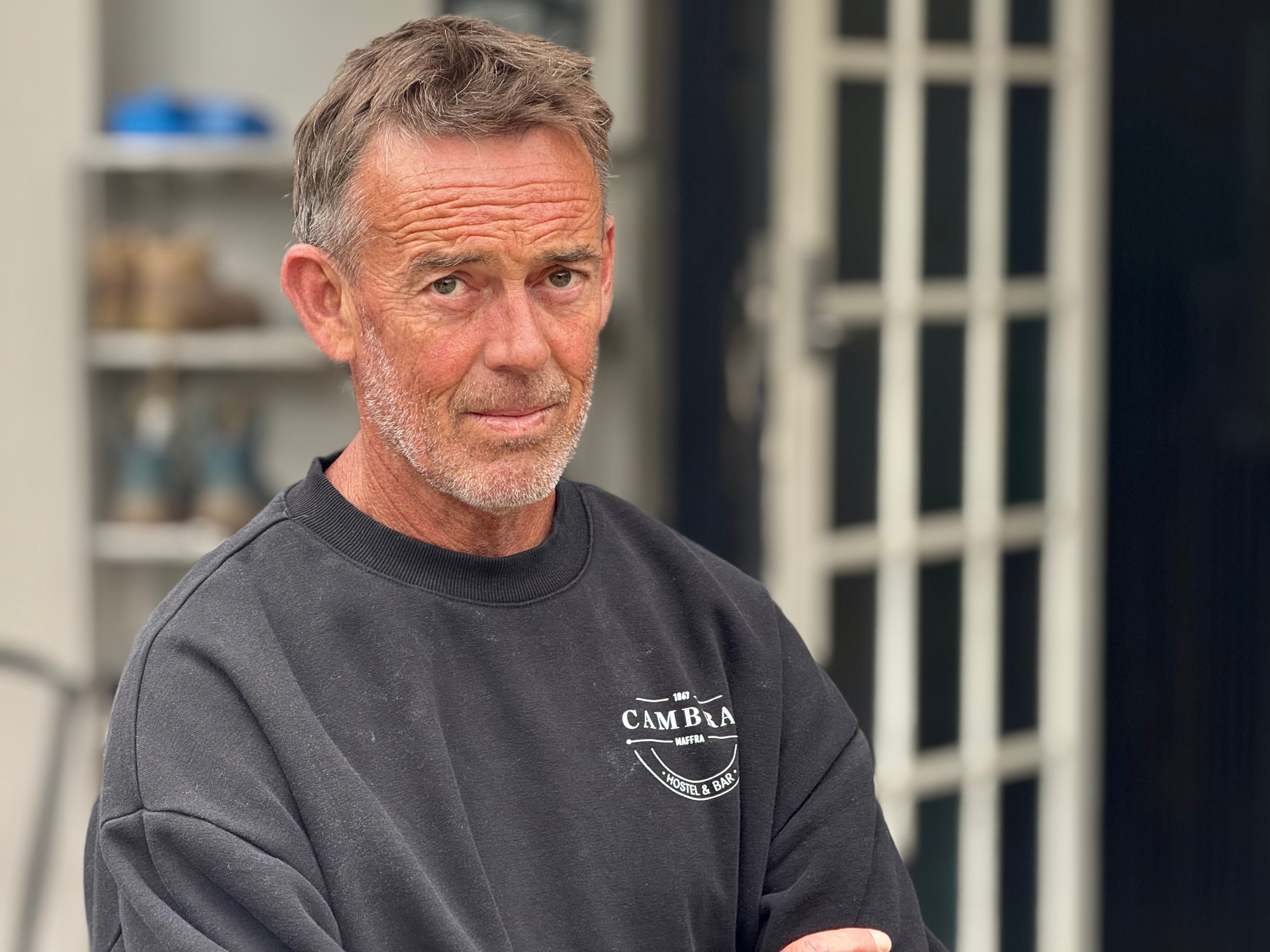 Un hombre de mediana edad, de cabello oscuro y vestido con un suéter oscuro, se encuentra cerca de una puerta.