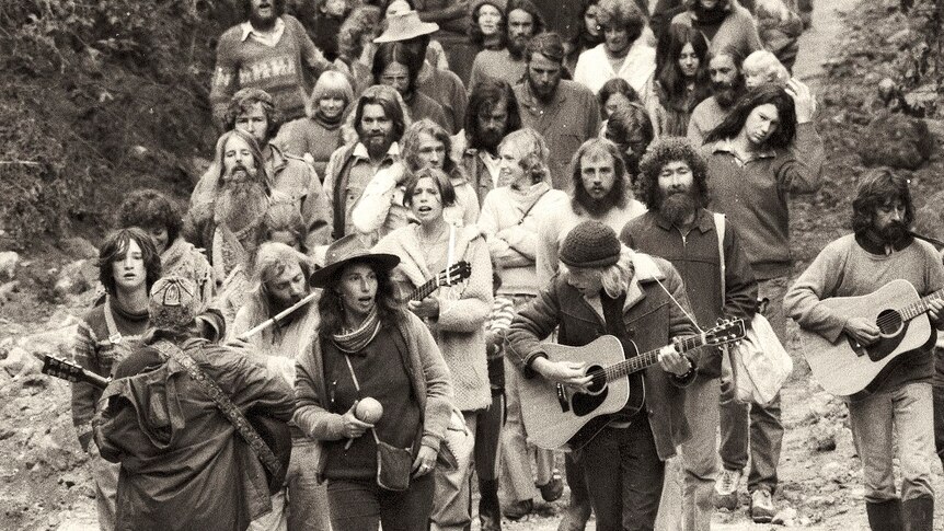 A black and white photo of about two dozen people walking down a road. The first two rows have instruments.