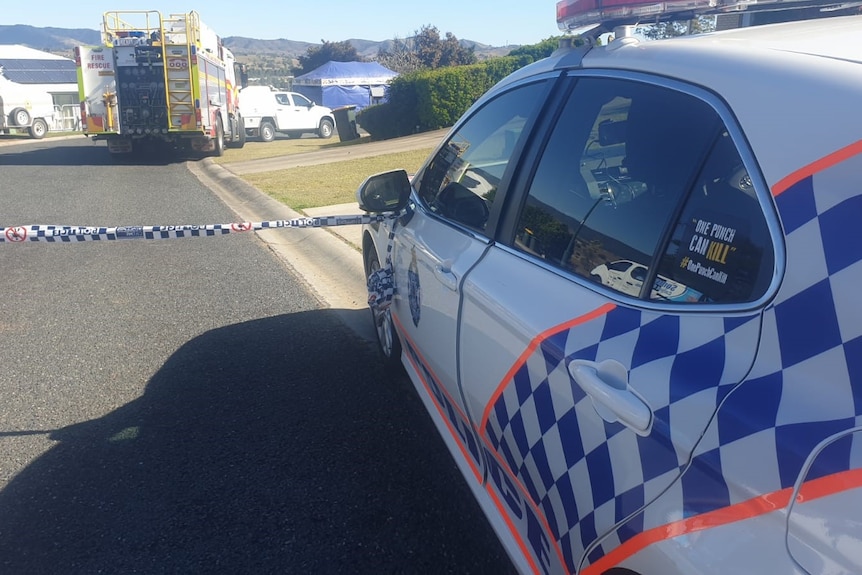 The side of a police car in front of police tape and a fire engine in the background.