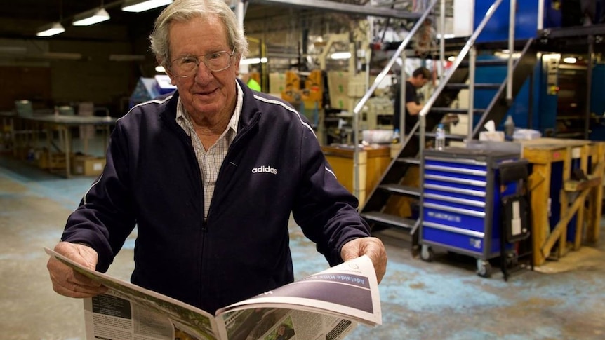 An elderly man stands in front of a printing plant, holding open a newspaper.