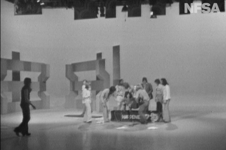 People gather around a mock coffin on a TV set.