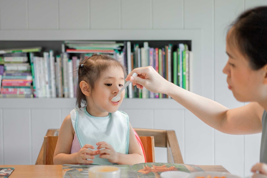 Woman feeding young child with a spoon sitting at a table.