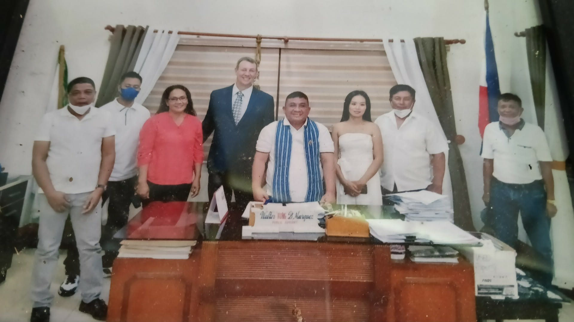 Photo of a photo with a bride and a groom standing in between priest with some other people standing behind a desk.