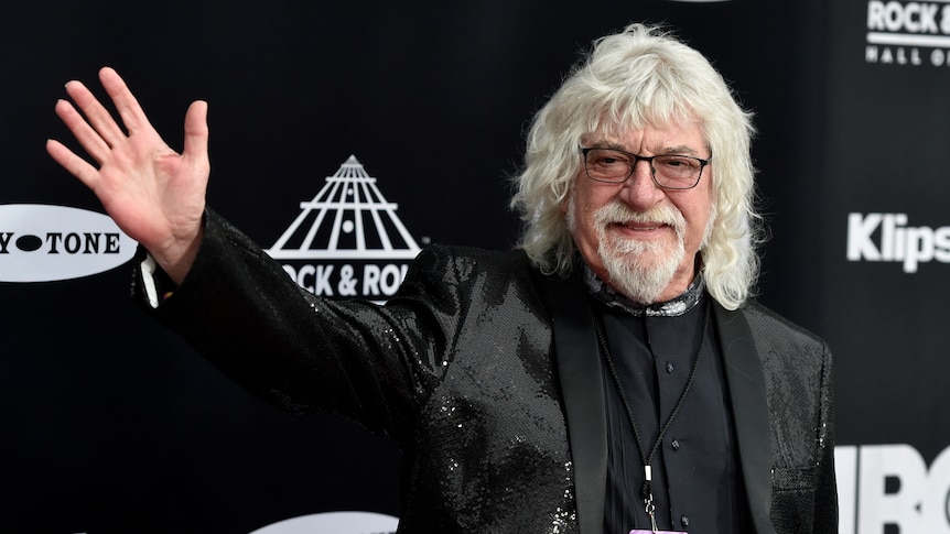 An older man with long white hair and glasses waves on the red carpet of an event.