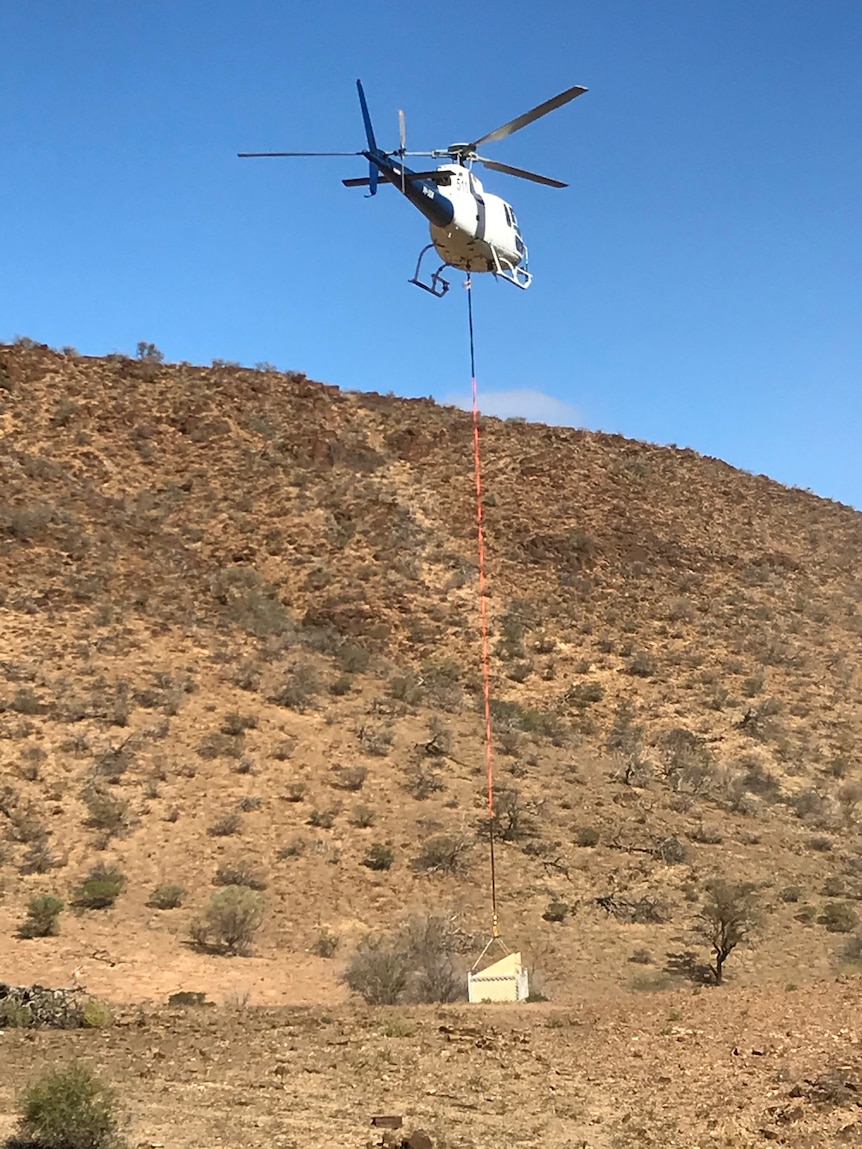 A helicopter towing a white box underneath it, against an arid mountainous landscape.