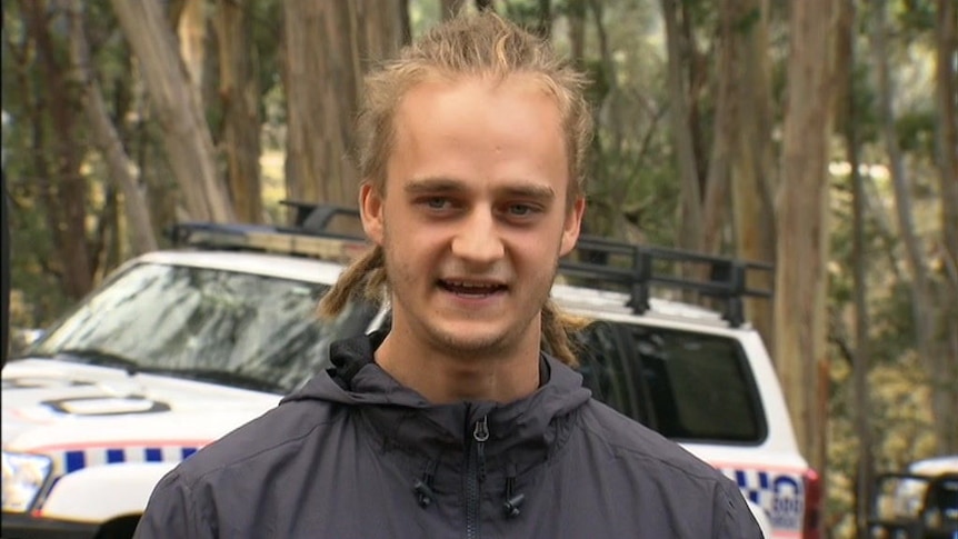 A man in a rainjacket speaks to the media in front of a police car in bushland.