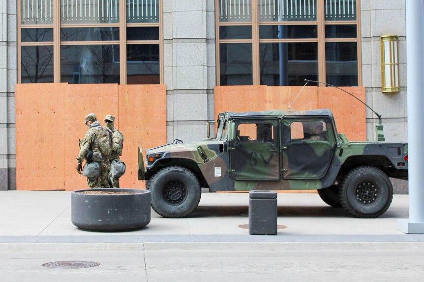 Two men in combat fatigues stand in front of a military vehicle outside a boarded-up building