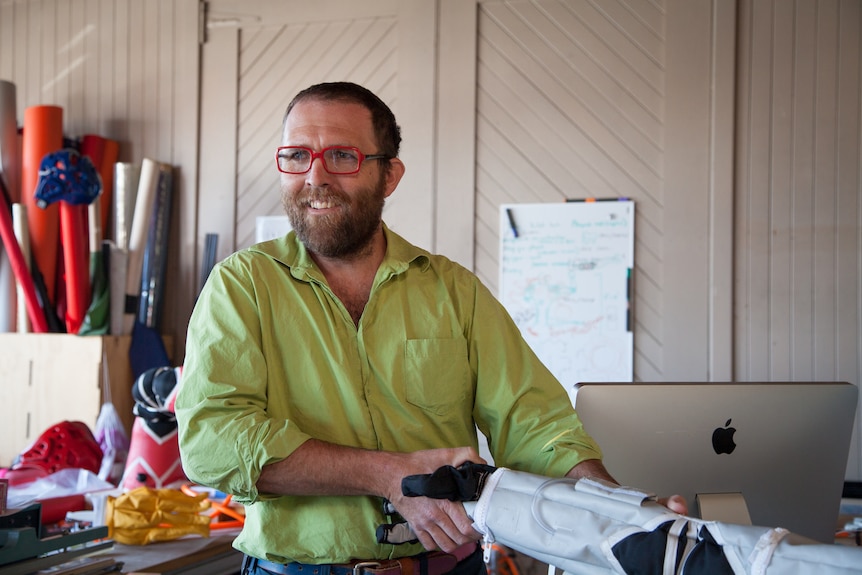 Man in a green button-down shirt and red glasses smiles, standing in a design lab, shaking hands with a robot arm