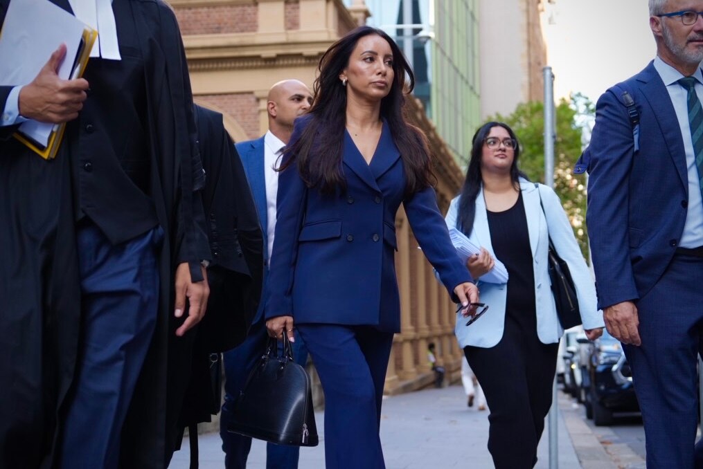 A woman wearing a blue paint suit walks towards a courthouse, flanked by people wearing suits and traditional lawyer attire