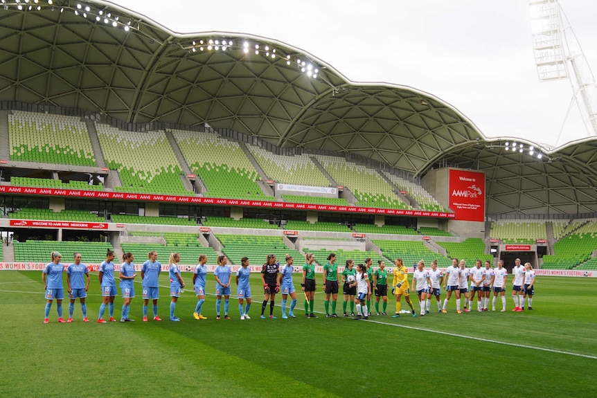 Melbourne City and Sydney FC W-League players line up alongside each other inside an empty stadium.