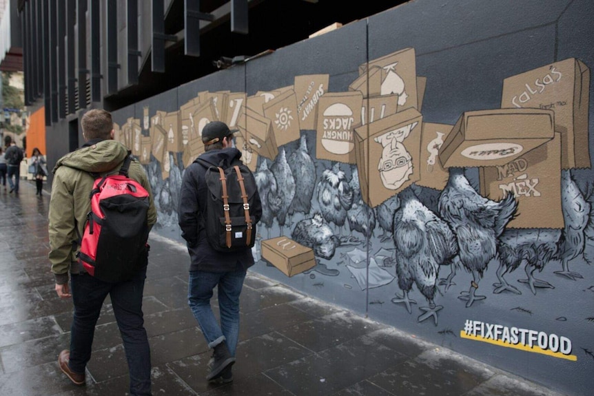 Two men walk past a mural in Melbourne that shows chickens with paper bags of fast food chains over their heads.