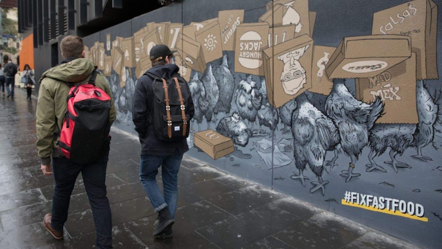 Two men walk past a mural in Melbourne that shows chickens with paper bags of fast food chains over their heads.