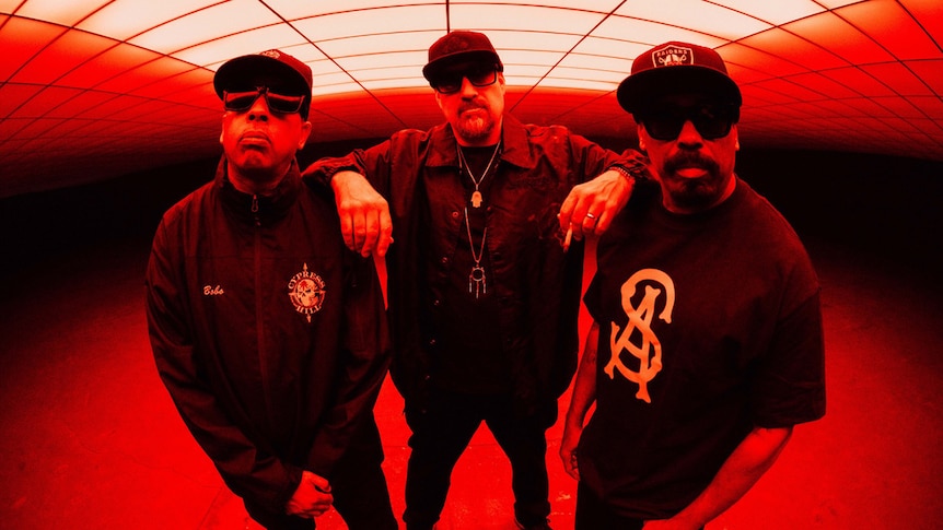 Three members of Cypress Hill stand in a red-lit room. They all wear sunglasses, B-Real holds a hand rolled cigarette or joint