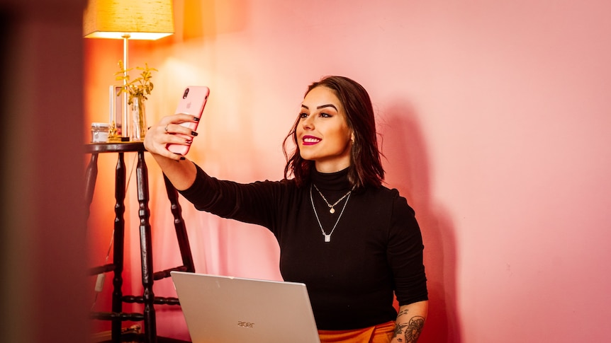 A woman with short hair in a black turtleneck shirt takes a selfie while sitting in front of a laptop