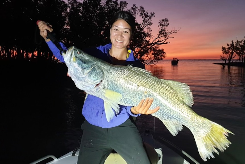 A woman, Loren Hanton, holding a large barramundi fish wearing a blue shirt at dusk in waters in the Northern Territory