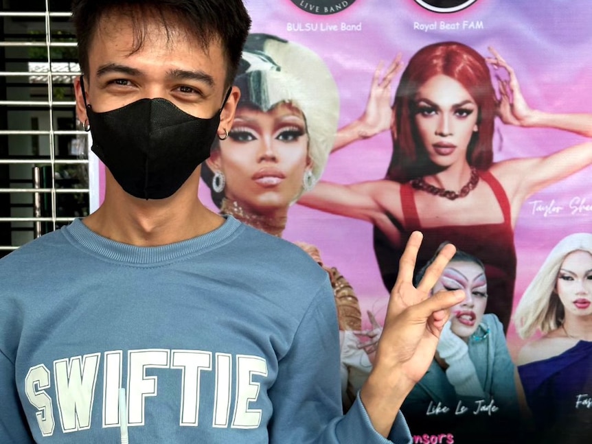 Mac smiles in a Swiftie jumper, wearing a mask and posing next to a poster which has a picture of him in drag on it.