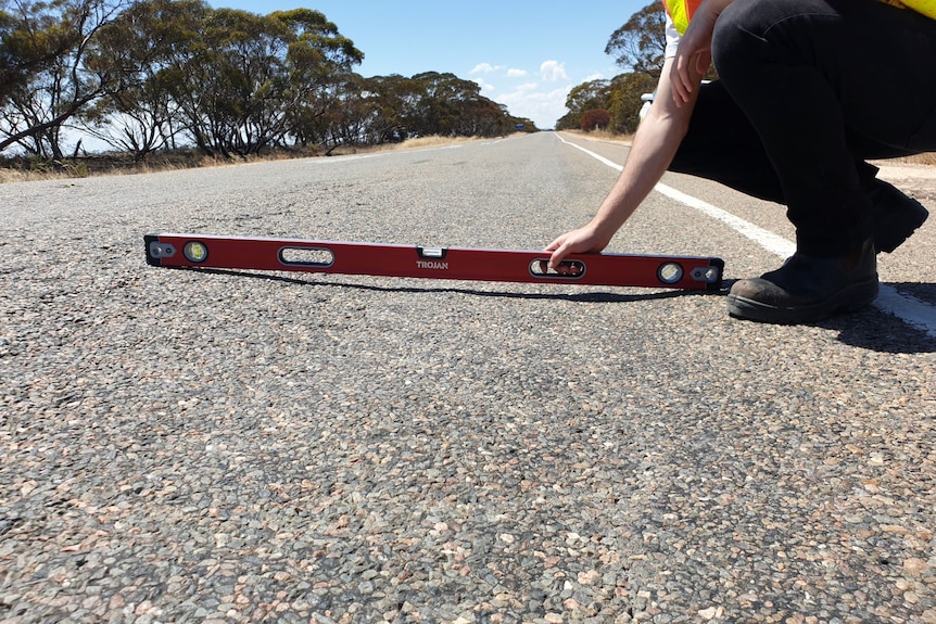 A man holding a spirit level against an uneven road. 