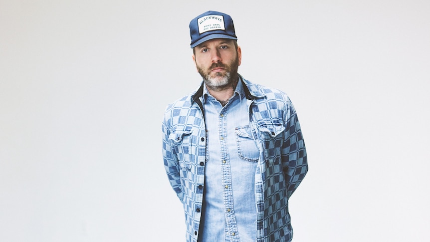 Dllas Green on City and Colour stands in a blank room with hands behind his back wearing a truckers cap.