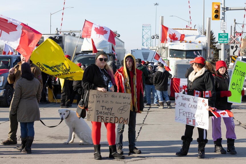 People with Canadian flags and protest signs stand on a road blocking traffic flanked by trucks.