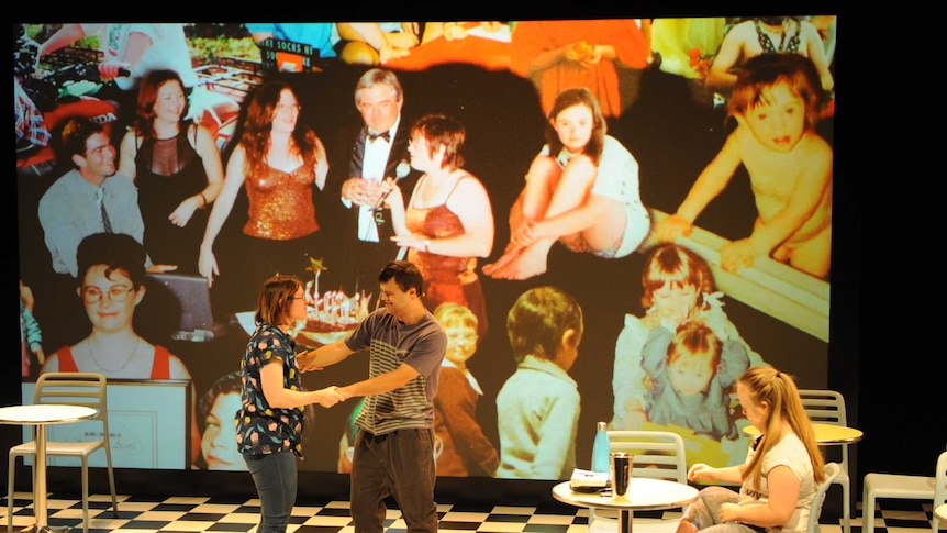 Three performers with Down Syndrome perform on stage with a background collage of childhood photos of the female performer.