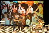 Three performers with Down Syndrome perform on stage with a background collage of childhood photos of the female performer.