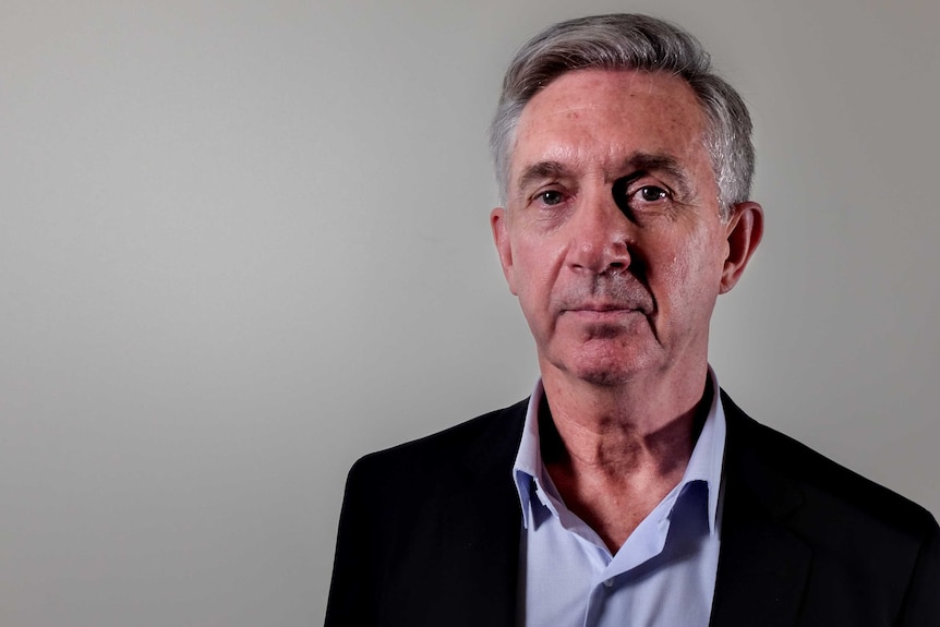 Ken Phillips, wearing a suit jacked and lilac shirt, stands in front of a grey backdrop.