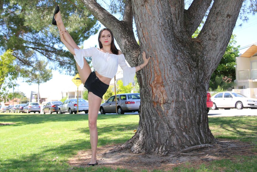 Vanessa stands with her leg above her head in a cheerleading pose with her hand on a tree.