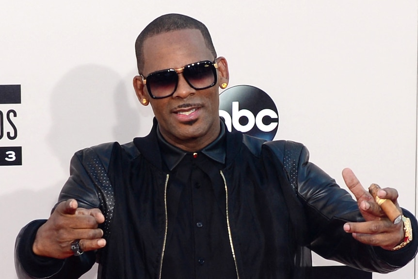 R Kelly wears sunglasses and a black jacket in front of a white NBC press screen.