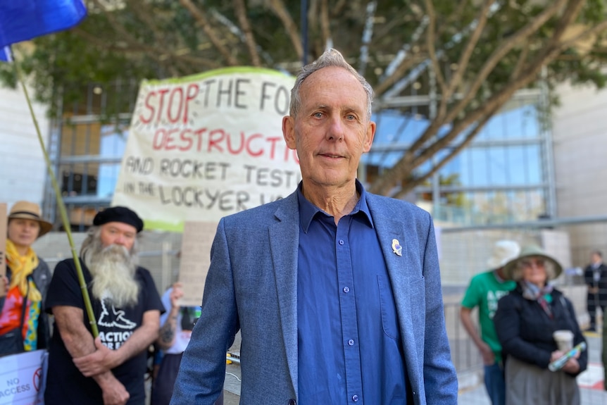 bob brown in a blue shirt and jacket with protesters behind him