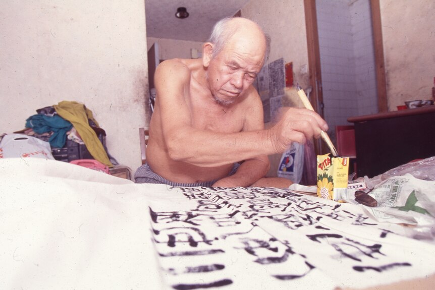 A shirtless older Hong Kong man sits in apartment and uses brush and black ink to write calligraphy.