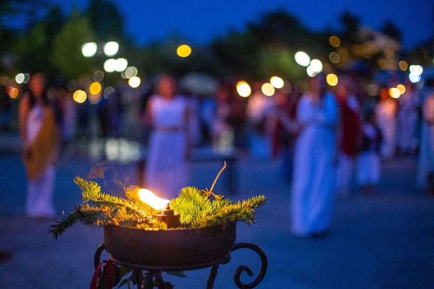 A candle burns in the foreground as women in ancient Greek dress mingle in the background.