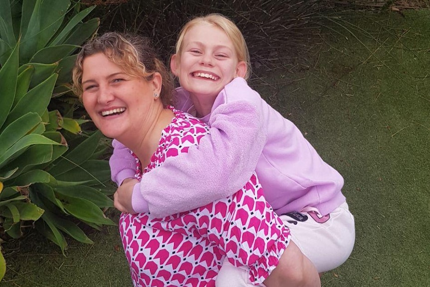 A woman in a pink shirt smiles up at the camera with her daughter hanging off her back. 