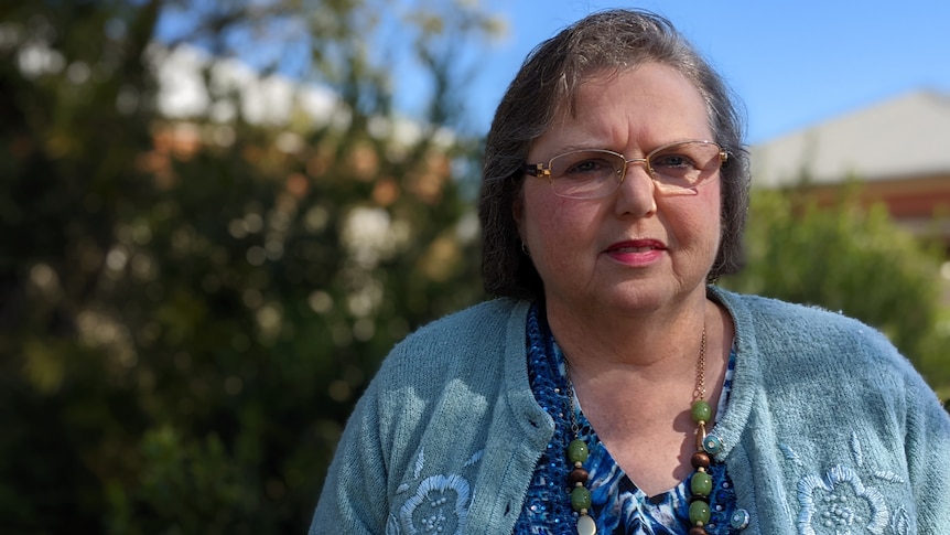 A close up of an older woman with glasses wearing a cardigan outside her home.