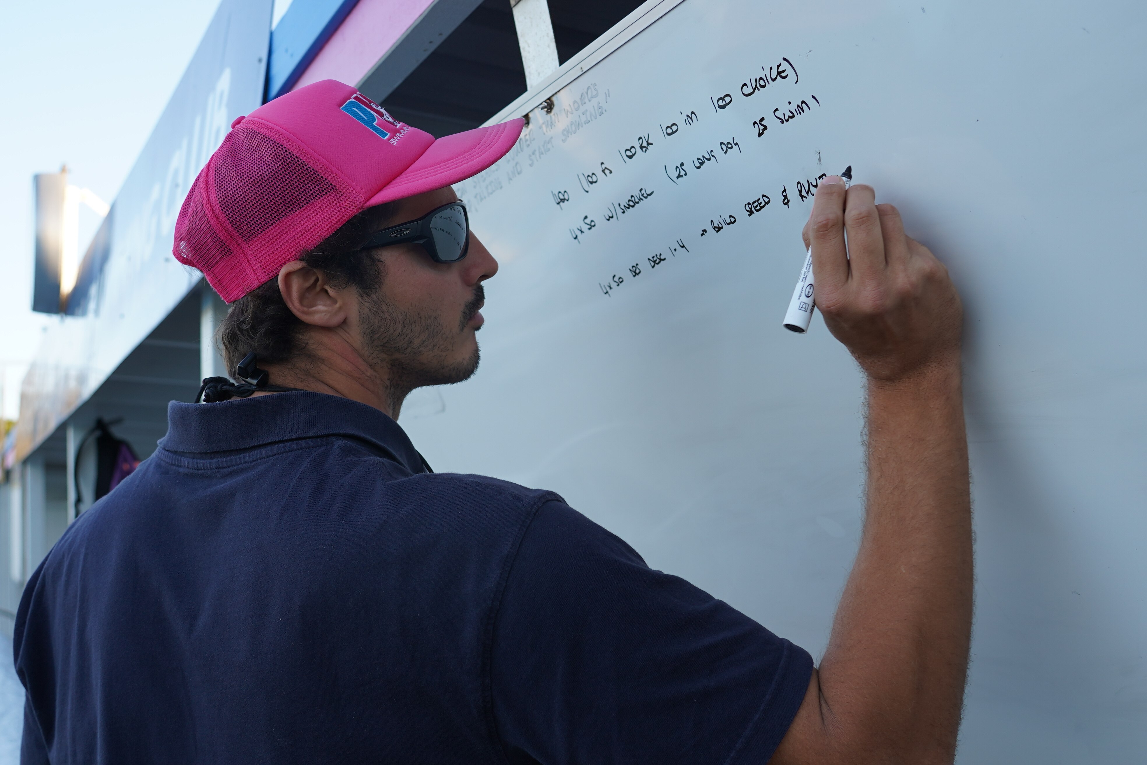 A man wearing a navy shirt, pink cap and sunglasses writes on a white board.