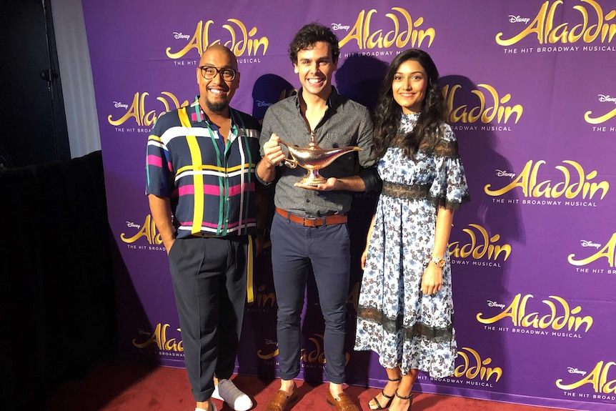 Actors Gareth Jacobs, Ainsley Melham and Shubshri Kandiah stand in front of an Aladdin backdrop.