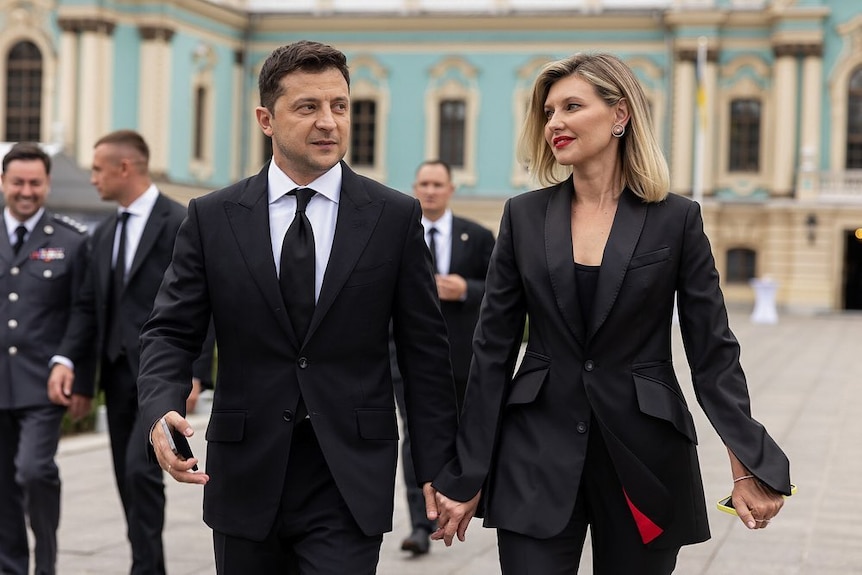 Volodymyr Zelenskyy and his wife Olena walk holding hands, both in black suits. hers has a red lining to match her bold lip