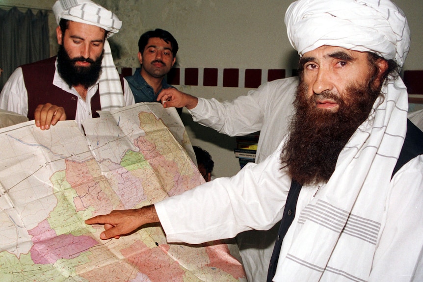 Jalaluddin Haqqani is seen with a long beard dressed in Afghan traditional clothes pointing at a map of Afghanistan.