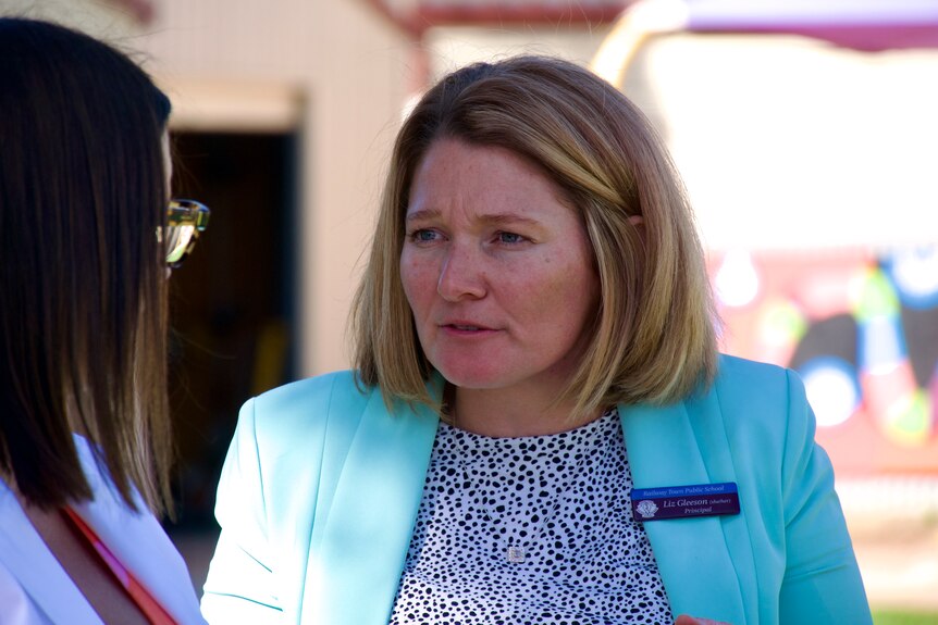 A blonde woman wearing a aqua cloloured blazer looking at a brown haired woman with glasses